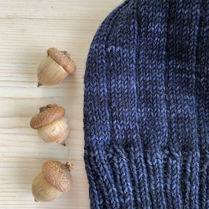 Puede incluir: Un gorro de punto azul oscuro con textura de canal&eacute;. Tres bellotas est&aacute;n dispuestas en fila sobre una superficie de madera clara.
