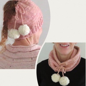 Könnte beinhalten: Eine rosa gestrickte Mütze mit einem Pferdeschwanzloch und zwei weißen Bommeln. Die Mütze ist hinten mit einem rosa Band gebunden. Ein passender rosa gestrickter Schal mit zwei weißen Bommeln ist ebenfalls abgebildet.