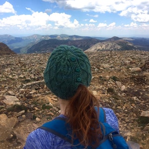 A woman is seen from behind standing on a rocky mountaintop with scenic views wearing a knitted hat with buttons, allowing her ponytail to stick out.