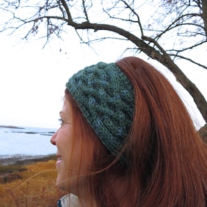 May include: A woman with long, red hair is wearing a green and blue knitted headband. She is looking out at a body of water.
