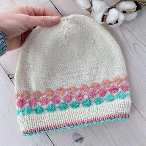 May include: Cream-colored knit beanie with a colorful geometric pattern. The pattern features rows of pink, peach, and teal diamond shapes. The brim and the edge of the pattern are trimmed in teal and pink.