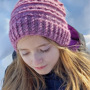 Puede incluir: Una persona joven lleva una gorra de punto morada con un patrón texturizado. La gorra está bajada sobre la frente y la persona mira hacia abajo. Lleva una chaqueta negra con un logotipo morado.