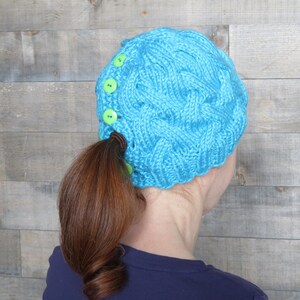 Peut inclure: Un bonnet en tricot bleu turquoise avec un trou pour la queue de cheval et des boutons verts sur le côté.