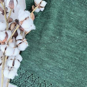 May include: A close-up of a green knitted top with a decorative scalloped edge. The top is made of a textured yarn and has a simple, round neckline. A sprig of cotton bolls is in the upper left corner.