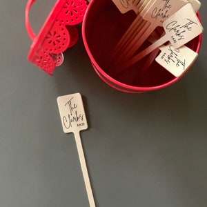 May include: A set of wooden stick place cards with the text "The Clarks" and the date "6.4.22" printed on them. The place cards are in a red bucket with a scalloped edge.