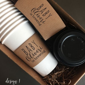 May include: A brown box filled with brown paper shreds contains a white coffee cup with a brown paper sleeve that reads "BABY Oliveri is brewing".  A black coffee lid is also in the box.