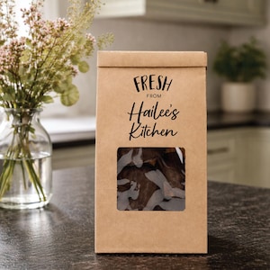 May include: A brown paper bag with a rectangular window, displaying a glimpse of the contents. The bag features the text "FRESH FROM Hailee's Kitchen" in black script. The bag is on a dark countertop, with a vase of flowers in the background.