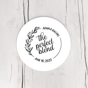 May include: A white, circular sticker with black text and a floral design. The text reads "Adam & Rachel" above "the perfect blend" and "June 10, 2022". The design includes a wreath of leaves and berries.