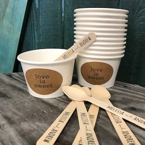 May include: White paper cups with brown paper circles that say "love is sweet" and wooden spoons with the text "Melissa and Andrew" printed on them.