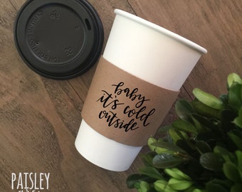 May include: A brown paper coffee cup sleeve with the text "baby it's cold outside" in black script. The sleeve is on a white coffee cup.