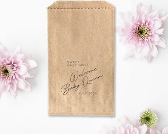 May include: A brown paper favor bag with the text "Sweet Baby Girl! Welcome Baby Quinn July 2024" in black script. The bag is surrounded by pink flowers with green centers, set against a white background.