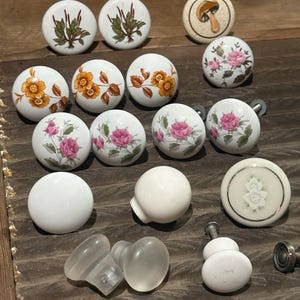 May include: A collection of round cabinet knobs featuring various designs including botanical plant illustrations, orange and pink floral patterns, a single mushroom graphic, solid white ceramic finishes, and translucent frosted glass options.