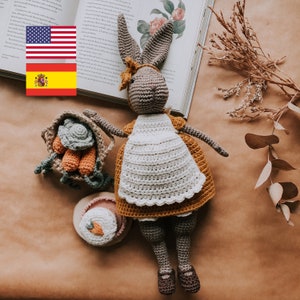 Könnte beinhalten: Ein gehäkeltes Hasenspielzeug, das eine weiße Schürze und ein gelbes Kleid trägt. Der Hase sitzt auf einer braunen Oberfläche mit einem kleinen gehäkelten Korb mit Karotten und einem kleinen gehäkelten Kuchen.