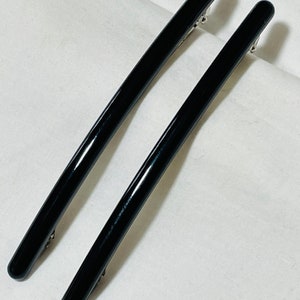 May include: Two black hair clips with a shiny finish. The clips are long and slender, with a slight curve.
