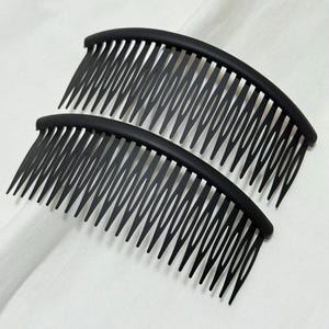May include: Two black hair combs with curved tops and evenly spaced teeth. The combs are made of a matte black material and are arranged side by side against a white background. The teeth are pointed and extend downwards.