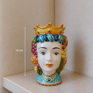 May include: A ceramic head vase, approximately 7 inches tall, featuring a woman's face with a crown and colorful floral and fruit details. The vase has a blue collar with a central jewel and is painted with vibrant colors.