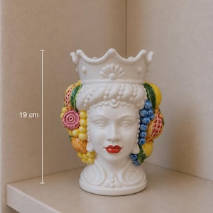 May include: A white ceramic vase, 19 cm tall, shaped like a woman's head with a crown. The head is adorned with colorful fruit and flowers. The woman has red lips and a detailed facial structure. The vase is a decorative item.