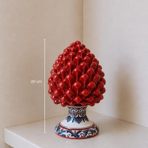 Op de afbeelding: Een levendige rode keramische dennenappelsulptuur, ongeveer 20 cm hoog, met een gedetailleerd, getextureerd oppervlak. De basis heeft een witte achtergrond met blauwe en rode bloempatronen, wat een decoratief tintje geeft.