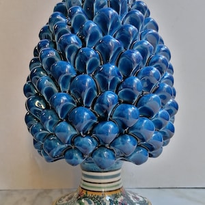 Puede incluir: Una escultura de piña de cerámica azul con una base detallada y estampada. El cono está hecho de muchas escamas individuales que se superponen.