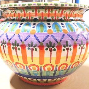 Może przedstawiać: Duży, okrągły, ceramiczny pojemnik na rośliny z kolorowym, ręcznie malowanym wzorem. Pojemnik zawiera r&oacute;żne kolory, w tym pomarańczowy, ż&oacute;łty, zielony, niebieski i fioletowy. Wz&oacute;r zawiera geometryczne kształty, paski i motywy kwiatowe.