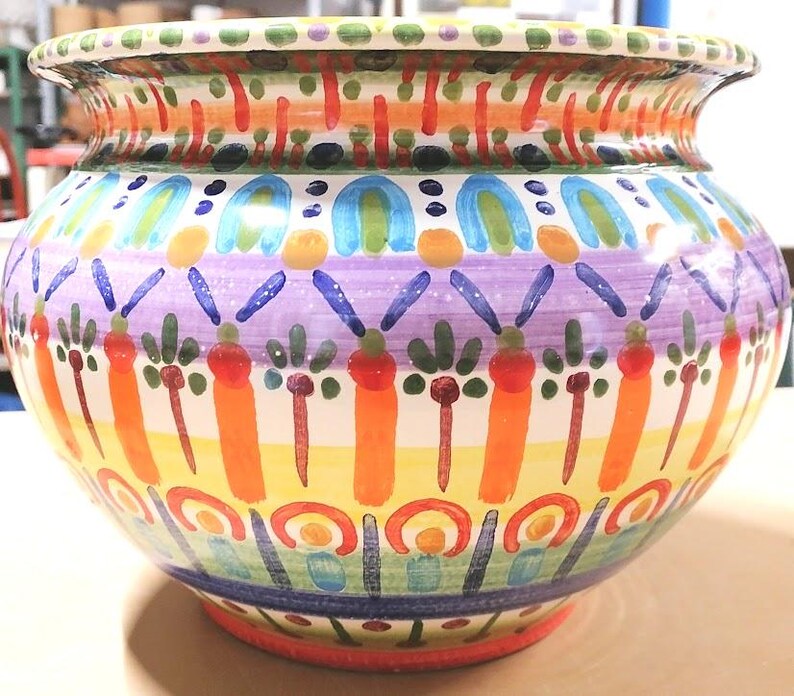 Może przedstawiać: Duży, okrągły, ceramiczny pojemnik na rośliny z kolorowym, ręcznie malowanym wzorem. Pojemnik zawiera r&oacute;żne kolory, w tym pomarańczowy, ż&oacute;łty, zielony, niebieski i fioletowy. Wz&oacute;r zawiera geometryczne kształty, paski i motywy kwiatowe.