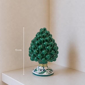 Może przedstawiać: Zielony ceramiczny przedmiot dekoracyjny w kształcie szyszki, o wysokości około 15 cm, z szczegółową, teksturowaną powierzchnią. Podstawa posiada malowany kwiatowy wzór z niebieskimi, żółtymi i zielonymi akcentami. Przedmiot jest wystawiony na białej półce.