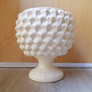 May include: White ceramic planter shaped like a pinecone with a pedestal base. The planter is a decorative piece for home decor.