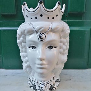 Könnte beinhalten: Weißer Keramiktopf in Form eines Frauenkopfes mit einer Krone, schwarzen und weißen Details und einem floralen Design um den Hals.