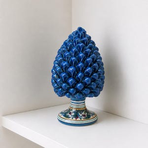Op de afbeelding: Een levendige blauwe keramische dennenappelsculptuur. De dennenappel is diep, glanzend blauw, met een textuur. Hij staat op een decoratieve basis met bloemen- en streepmotieven. Een uniek woondecoratie-item.