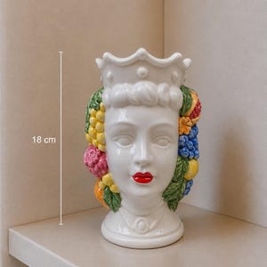 Op de afbeelding: Een witte keramische vaas van 18 cm hoog in de vorm van een vrouwenhoofd, versierd met een kroon en kleurrijk fruit. De vaas heeft rode lippen en gedetailleerde gelaatstrekken. Het fruit omvat druiven, citroenen en granaatappels.
