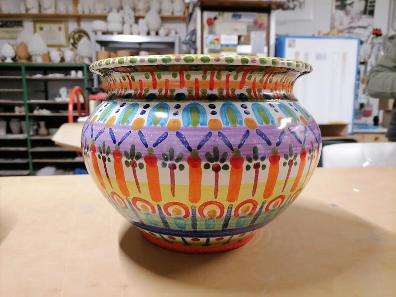 Może przedstawiać: Duży, okrągły, ceramiczny pojemnik na rośliny z kolorowym, ręcznie malowanym wzorem. Pojemnik zawiera r&oacute;żne kolory, w tym pomarańczowy, ż&oacute;łty, zielony, niebieski i fioletowy. Wz&oacute;r zawiera geometryczne kształty, kwiaty i liście.