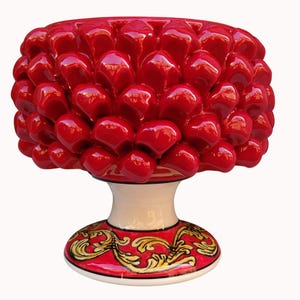 Puede incluir: Un objeto decorativo de cerámica con forma de piña y acabado en rojo brillante. La parte superior presenta escamas redondeadas y superpuestas. La base es un pedestal blanco con un patrón de volutas pintadas en rojo y dorado.
