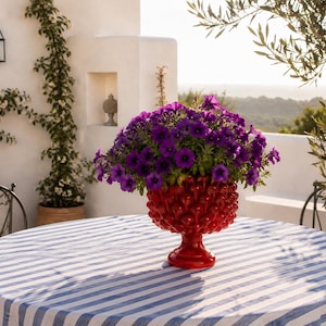 Puede incluir: Una mesa redonda al aire libre con un mantel a rayas azules y blancas. Un jarrón rojo texturizado lleno de flores moradas está en el centro. La escena está ambientada en un patio con plantas trepadoras y vistas al paisaje.