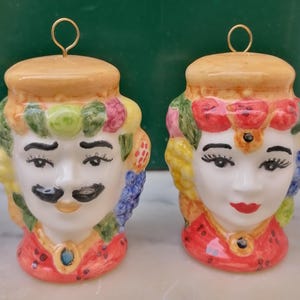 May include: Two ceramic figurines, one male and one female, with colourful, ornate designs. The male figure has a black moustache, while the female has red lips. Both have a gold loop at the top for hanging.
