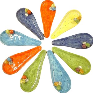 Può includere: Un set di dieci cucchiai in ceramica di vari colori, ognuno con un piccolo disegno floreale. I cucchiai sono disposti in cerchio, con le punte rivolte verso l'esterno.