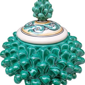 Pode incluir: Um pote de cerâmica verde com uma tampa em forma de pinha. O pote é decorado com padrões florais verdes, amarelos e azuis.