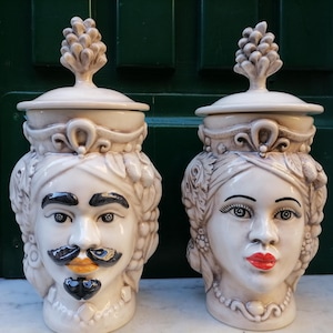 May include: Two ceramic jars with lids, each shaped like a human head. One has a mustache and goatee, the other has red lipstick. Both are off-white with detailed facial features and a decorative top.