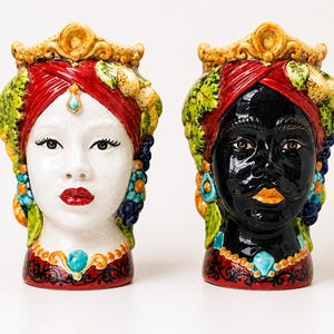 Puede incluir: Dos esculturas de cabeza de cerámica, una con rostro blanco y otra con rostro negro. Ambas tienen pañuelos rojos, coronas doradas y detalles coloridos en verde, azul y amarillo. Las esculturas son decorativas y artísticas.