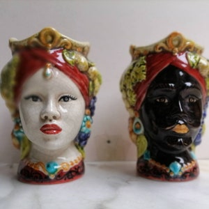 Könnte beinhalten: Zwei Keramikkopfskulpturen. Eine ist eine hellhäutige Frau mit roten Lippen, einem roten Kopftuch und einer juwelenbesetzten Krone. Die andere ist eine Person mit schwarzer Haut mit Schnurrbart, rotem Kopftuch und juwelenbesetzter Krone. Beide haben verzierte Details.