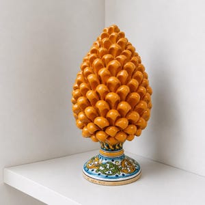 Può includere: Una scultura decorativa a forma di pigna in ceramica arancione. La pigna ha una superficie strutturata e poggia su una base dettagliata con motivi blu, verdi e gialli. La scultura è esposta su una mensola bianca.