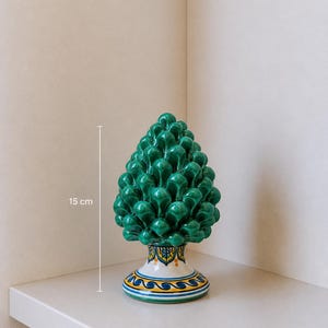 May include: A green ceramic pinecone ornament, about 15 cm tall, with a detailed, textured surface. The base is white with blue, yellow, and gold decorative patterns. It is displayed on a white shelf against a neutral background.