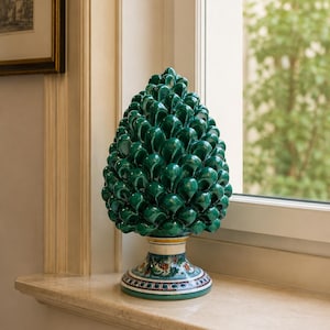 Pode incluir: Uma escultura decorativa de pinha de cerâmica verde brilhante. A pinha assenta sobre uma base detalhada com desenhos florais e geométricos. A escultura é exibida numa soleira de mármore.