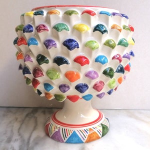 May include: A white ceramic vase with a textured surface, adorned with colorful, raised, rounded shapes. The vase has a pedestal base with a red band and geometric patterns. The rim is red.