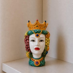 May include: A ceramic head vase with a white face, red lips, and a golden crown. The head is adorned with colorful fruit and jewels. The vase is a decorative item, likely for home decor.