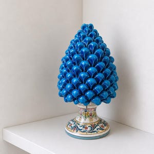 Könnte beinhalten: Eine leuchtend blaue Keramik-Kiefernzapfenskulptur. Der Kiefernzapfen ist ein tiefes, glänzendes Blau und steht auf einem dekorativen Sockel mit bunten Blumenmustern. Die Skulptur wird auf einem weißen Regal ausgestellt.