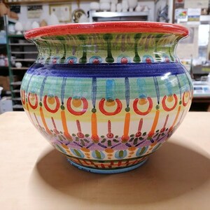 Może przedstawiać: Kolorowa doniczka ceramiczna z geometrycznym wzorem w odcieniach niebieskiego, zielonego, ż&oacute;łtego, pomarańczowego i czerwonego. Doniczka ma szeroką, okrągłą podstawę i lekko rozszerzający się rant.