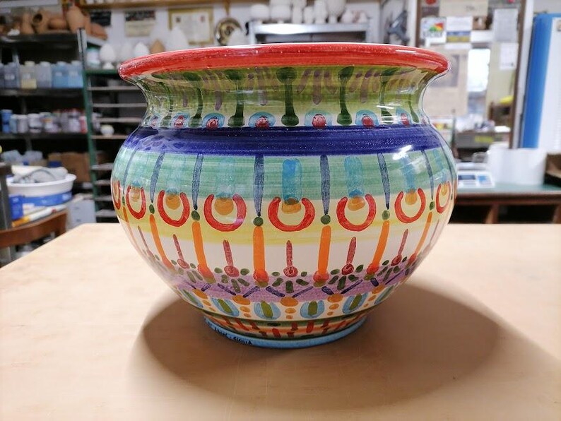 Może przedstawiać: Kolorowa doniczka ceramiczna z geometrycznym wzorem w odcieniach niebieskiego, zielonego, ż&oacute;łtego, pomarańczowego i czerwonego. Doniczka ma szeroką, okrągłą podstawę i lekko rozszerzający się rant.