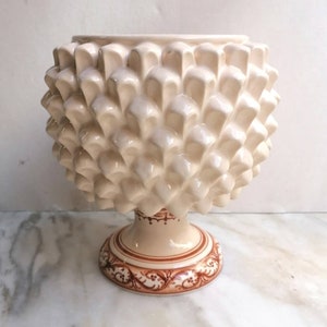 Pode incluir: Um vaso de cerâmica branco em forma de pinha com uma base com padrão marrom e branco.