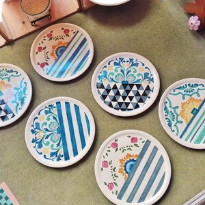 May include: Set of six decorative ceramic plates with various colorful patterns. The plates feature geometric designs, floral motifs, and stripes in shades of blue, green, yellow, and white.