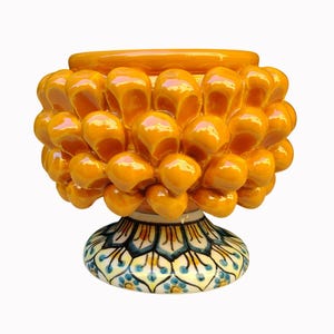 Pode incluir: Um vaso de cerâmica com uma secção superior texturizada de cor laranja brilhante que lembra uma pinha, composta por protuberâncias arredondadas. A base é um pedestal alargado decorado com um padrão simétrico detalhado em azul, amarelo e preto.